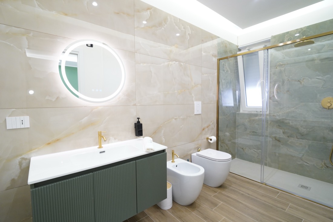 Bagno con mobile scanalato verde, specchio tondo retroilluminato e rubinetteria dorata